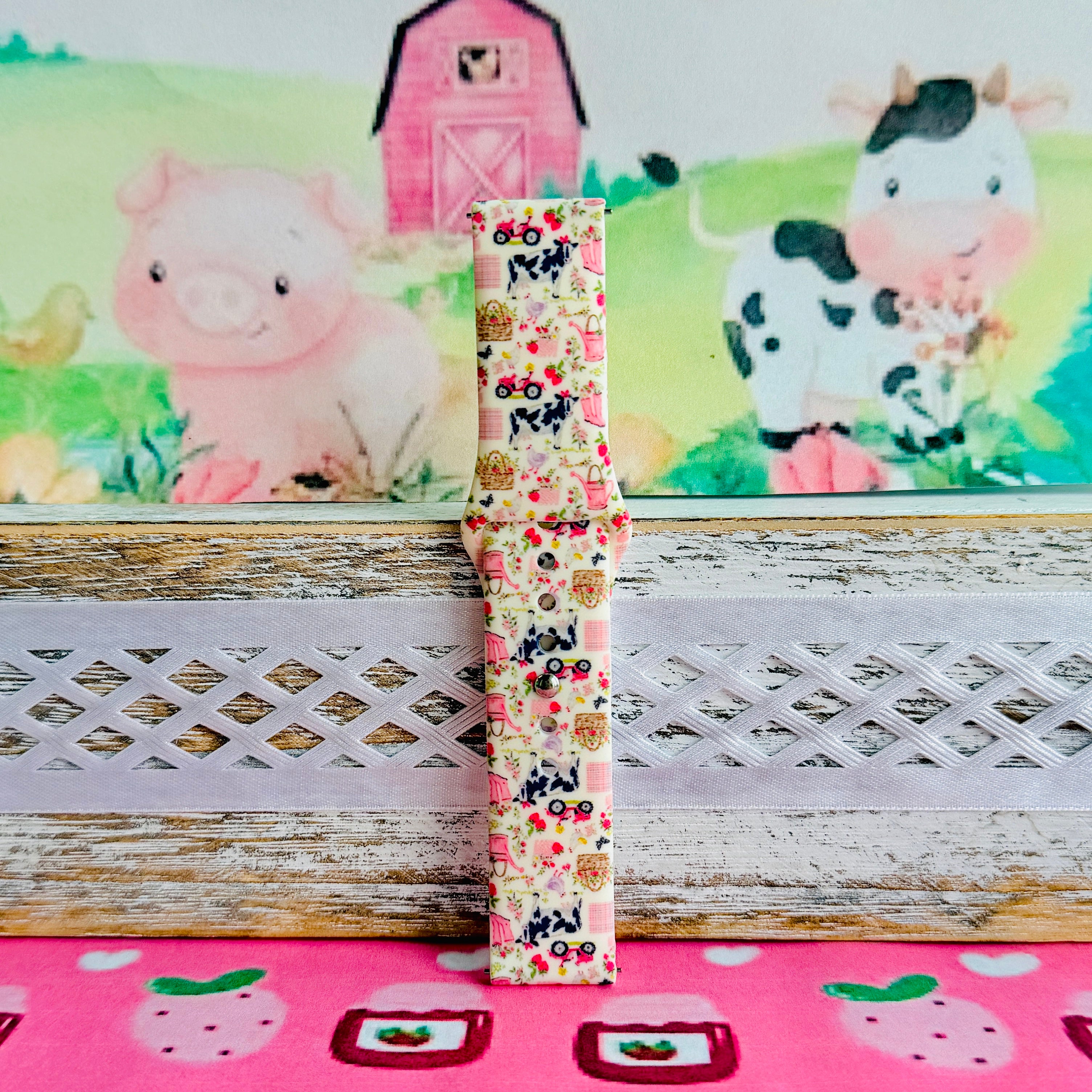 Farm Girl Cutie silicone Samsung Galaxy Watch band with dairy cows, red tractors, chickens, ducks, pink watering cans, strawberries, baskets, and roses on a cream base with pink gingham squares, shown displayed and worn against a farmhouse country backdrop.