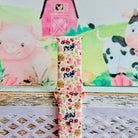 Farm Girl Cutie silicone Samsung Galaxy Watch band with dairy cows, red tractors, chickens, ducks, pink watering cans, strawberries, baskets, and roses on a cream base with pink gingham squares, shown displayed and worn against a farmhouse country backdrop.