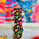 Bloom Bonita silicone Apple Watch band with Mexican folk art florals in coral, red, yellow, teal, and pink on a black base, shown displayed against a vibrant Mexican folk art backdrop with rose and dress accents.