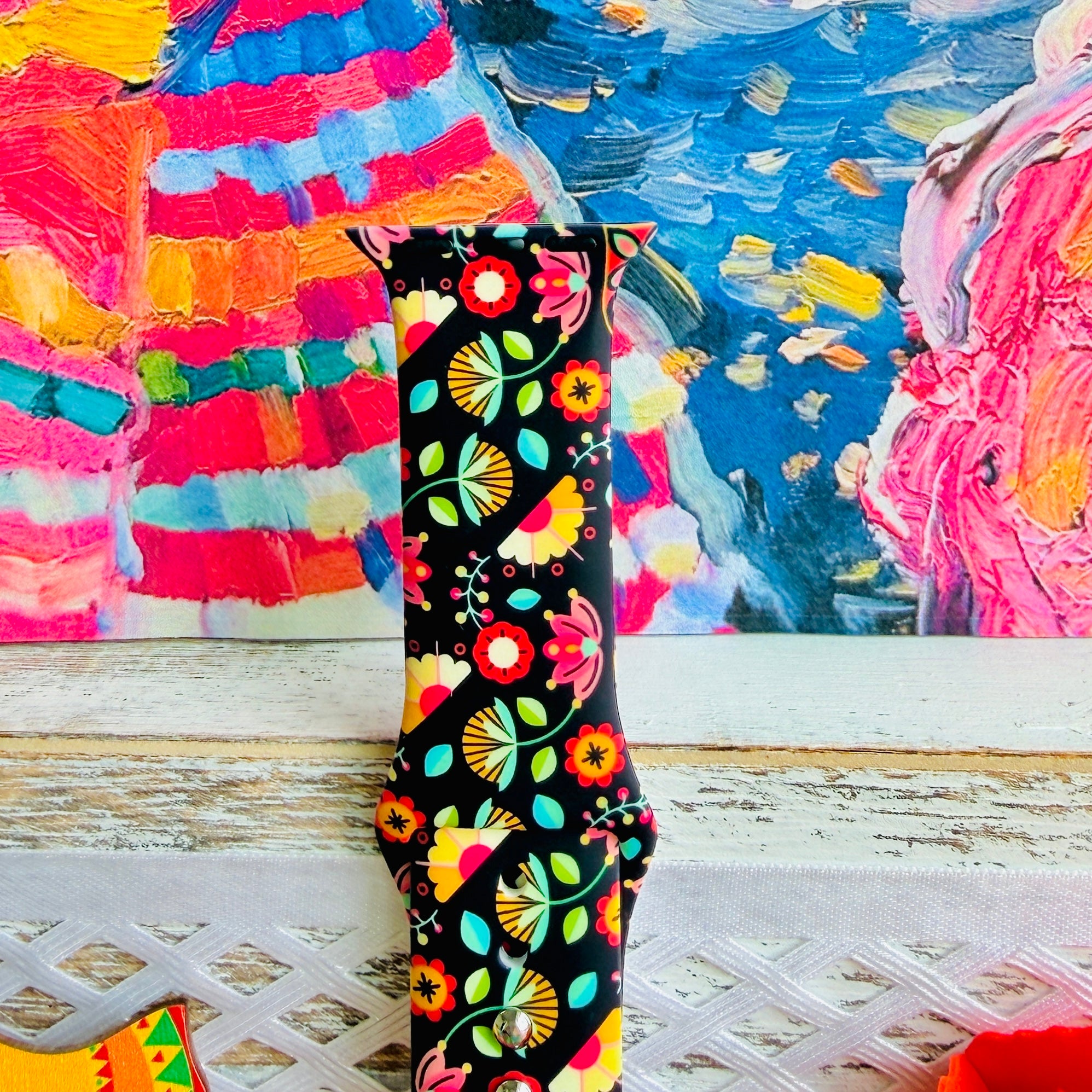 Bloom Bonita silicone Apple Watch band with Mexican folk art florals in coral, red, yellow, teal, and pink on a black base, shown displayed against a vibrant Mexican folk art backdrop with rose and dress accents.