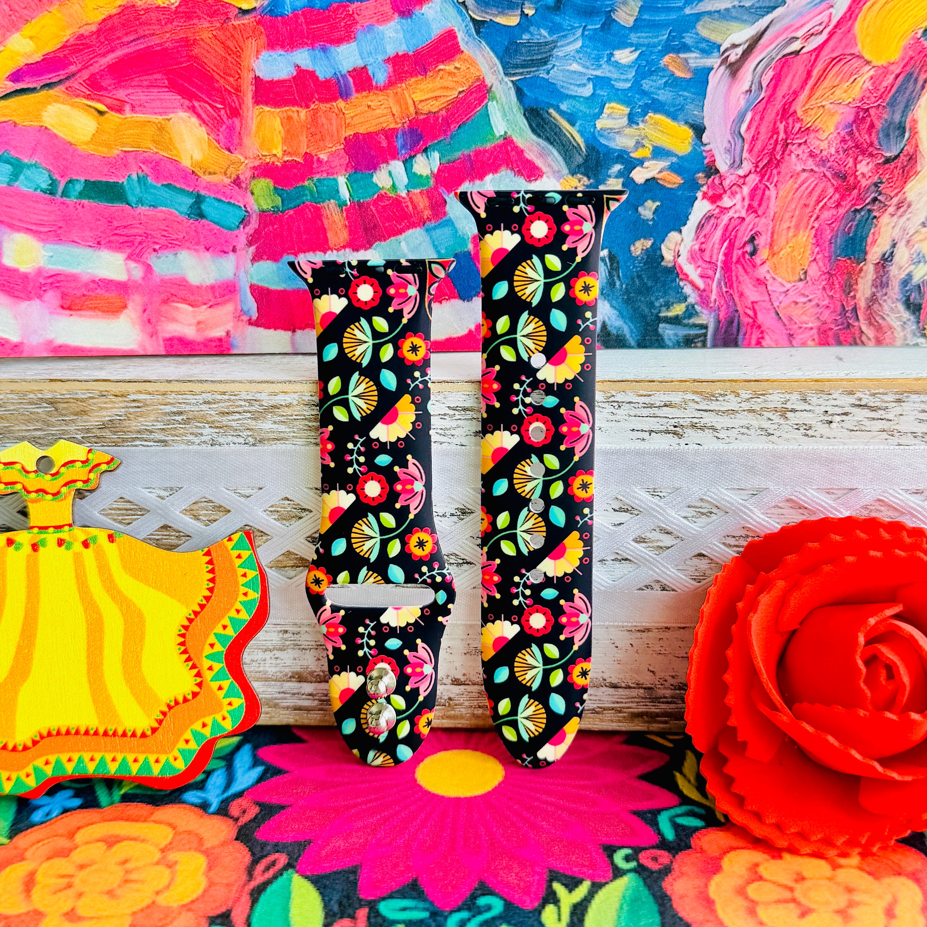 Bloom Bonita silicone Apple Watch band with Mexican folk art florals in coral, red, yellow, teal, and pink on a black base, shown displayed against a vibrant Mexican folk art backdrop with rose and dress accents.