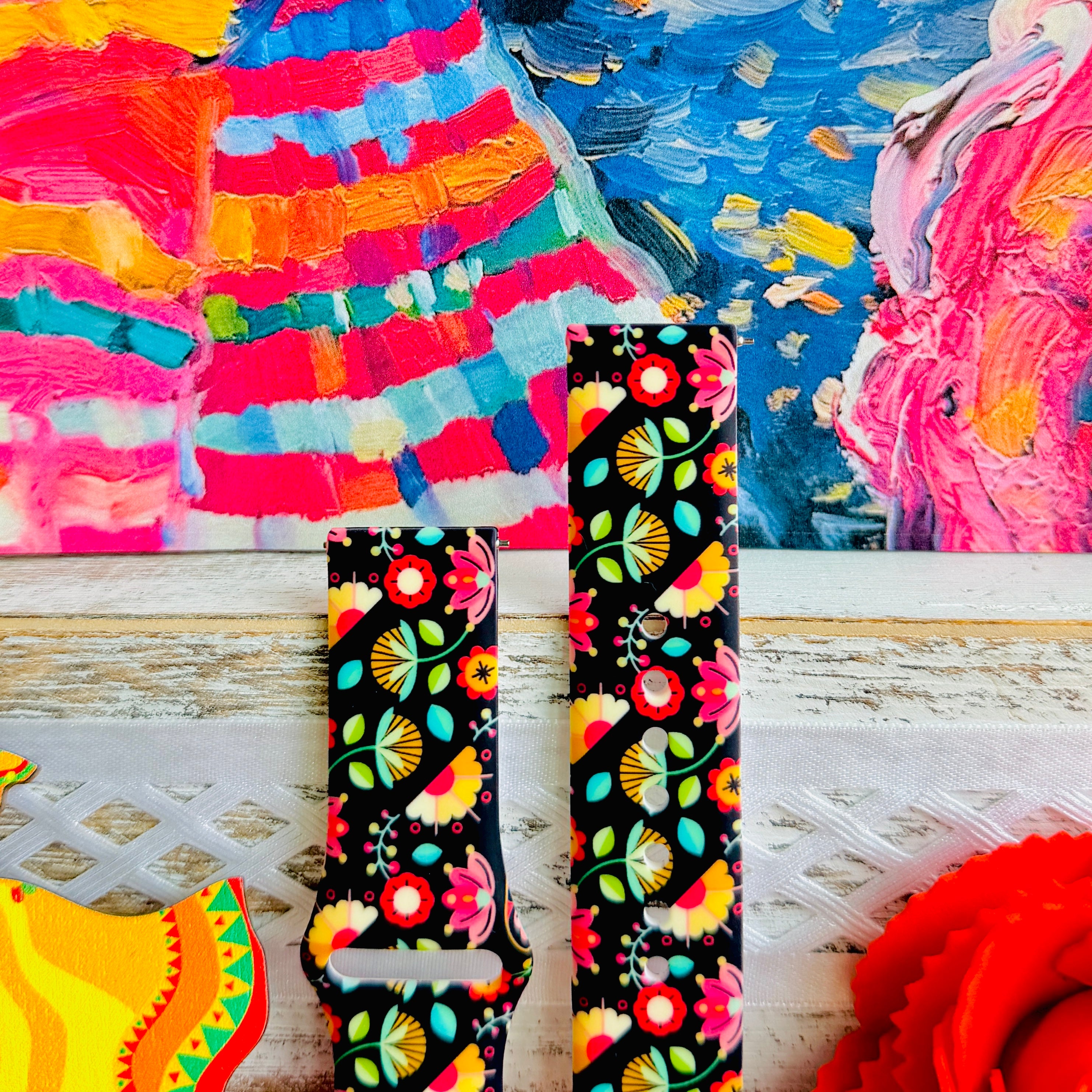Bloom Bonita silicone Samsung Galaxy Watch band with Mexican folk art florals in coral, red, yellow, teal, and pink on a black base, shown displayed against a vibrant Mexican folk art backdrop with rose and dress accents.