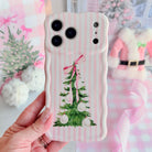 Hand holding the Pretty in PinkMas Wavy Luxe MagSafe iPhone case, a watercolor Christmas tree with pink bow and pearl ornaments on pink stripes, beside a pink Santa coat