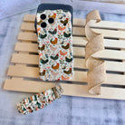 Cream and floral Cluckin' Cute scrunchie phone wrist strap with a hand-illustrated chicken and wildflower print, clipped to the matching farmhouse chicken Wavy Luxe iPhone case, styled on a rustic wood and burlap background.