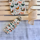 Cream and floral Cluckin' Cute scrunchie phone wrist strap with a hand-illustrated chicken and wildflower print, clipped to the matching farmhouse chicken Wavy Luxe iPhone case, styled on a rustic wood and burlap background.