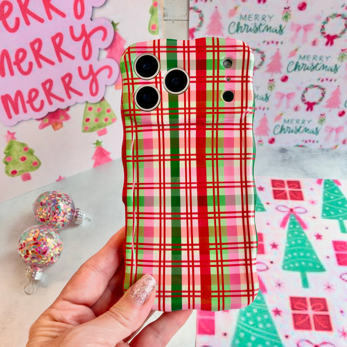 Hand holding the Merry Little Plaid Wavy Luxe MagSafe iPhone case in red, green and pink Christmas plaid, beside a pink Merry Christmas sign and glitter ornaments