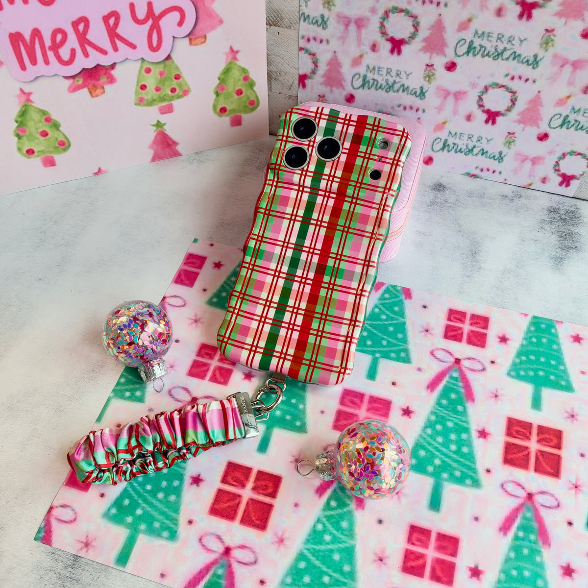 Merry Little Plaid iPhone case with the matching scrunchie phone wrist strap attached, laid flat on pink and mint Christmas paper