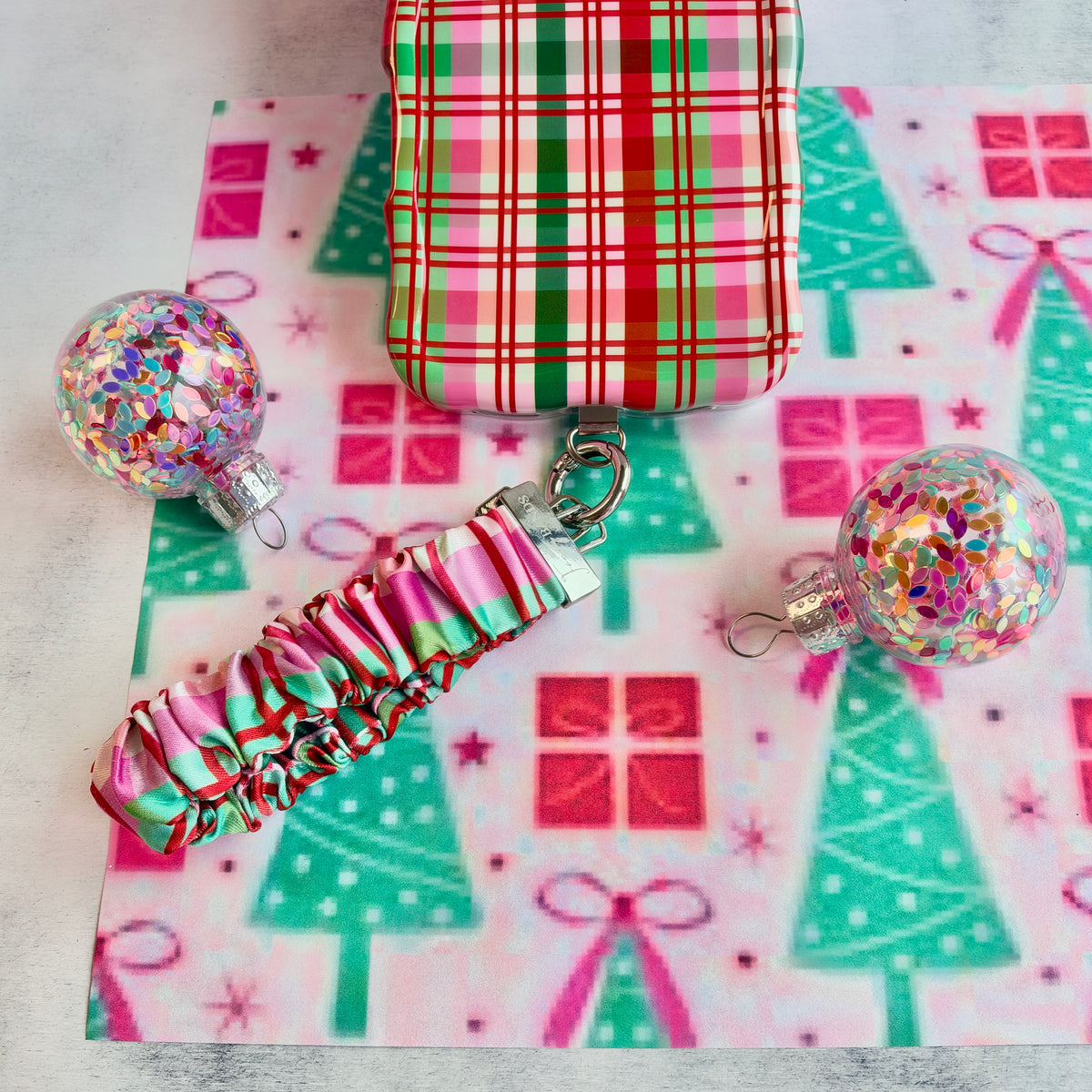 Merry Little Plaid scrunchie phone wrist strap clipped to the matching iPhone case, with glitter ornaments on Christmas tree paper