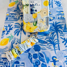 Blue and yellow La Dolce Summer scrunchie phone wrist strap in gingham and lemon print, clipped to the matching Amalfi Coast Wavy Luxe iPhone case with Italia and Capri travel stamps, styled on a blue-and-yellow Mediterranean palm and seashell print background.