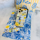 Blue and yellow La Dolce Summer scrunchie phone wrist strap in gingham and lemon print, clipped to the matching Amalfi Coast Wavy Luxe iPhone case with Italia and Capri travel stamps, styled on a blue-and-yellow Mediterranean palm and seashell print background.
