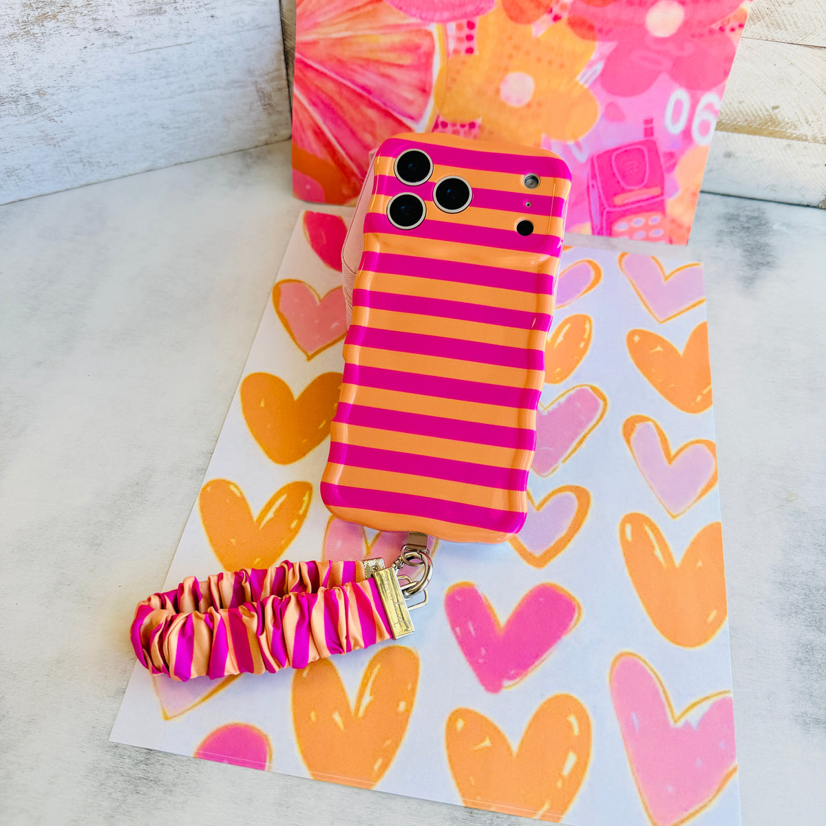 Candy Citrus Stripes scrunchie phone wrist strap laid out with the matching striped Wavy Luxe iPhone case on a heart print backdrop