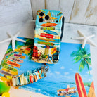 A colorful Seaside State Of Mind scrunchie phone wrist strap in a vintage beach print with Adirondack chairs, surfboard signposts, and palm trees, clipped to the matching Seaside State Of Mind Wavy Luxe iPhone case, styled on a tropical beach background with starfish and shells.