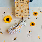 A sweet Bee Happy scrunchie phone wrist strap in a white bumblebee, honeycomb, and honey-pot print, clipped to the matching Bee Happy Wavy Luxe iPhone case, styled with sunflowers and bees on a soft cream background.
