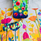 A vibrant Petal Princess scrunchie phone wrist strap in a colorful watercolor wildflower print, clipped to the matching Petal Princess Wavy Luxe iPhone case, styled on a bright painted floral background.