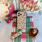 Flat lay of the Boot Scootin' Christmas iPhone case with its cowboy boot scrunchie wrist strap, a pink stocking and cowboy hat ornament
