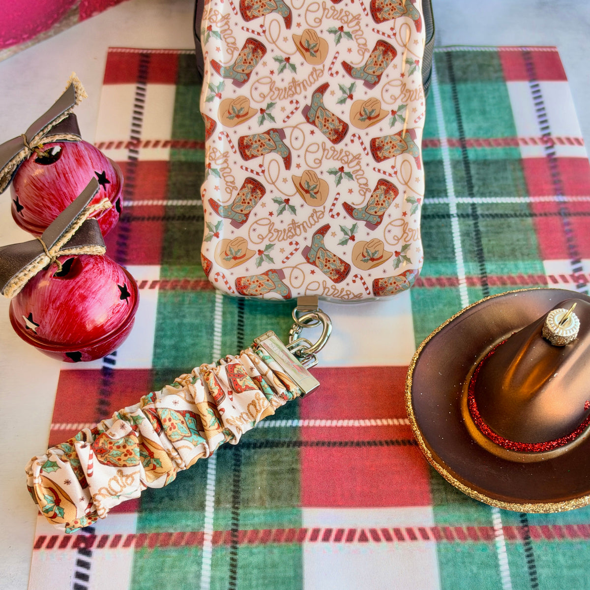 Boot Scootin' Christmas scrunchie phone wrist strap clipped below the matching iPhone case on plaid, with red jingle bells and a cowboy hat ornament