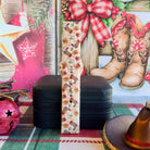 Boot Scootin' Christmas Samsung Galaxy Watch band buckled and standing on a black jewelry box between a jingle bell and a cowboy hat ornament