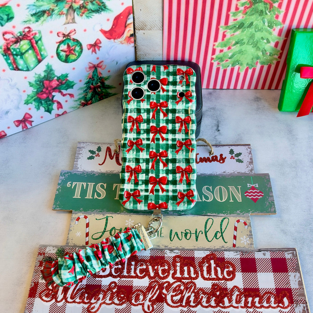 Plaid Tidings & Bows iPhone case and gingham bow scrunchie wrist strap on stacked wooden Christmas signs with a green gift box