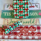 Plaid Tidings & Bows scrunchie phone wrist strap clipped below the matching iPhone case on Tis the Season and Believe in the Magic of Christmas signs