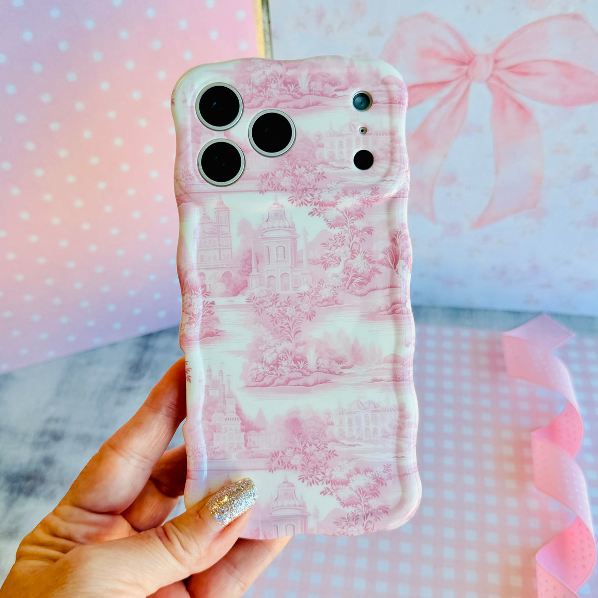 Hand holding the Blush Manor Wavy Luxe MagSafe iPhone case, pink toile manor houses and gardens on a cream ground, against a pink bow backdrop