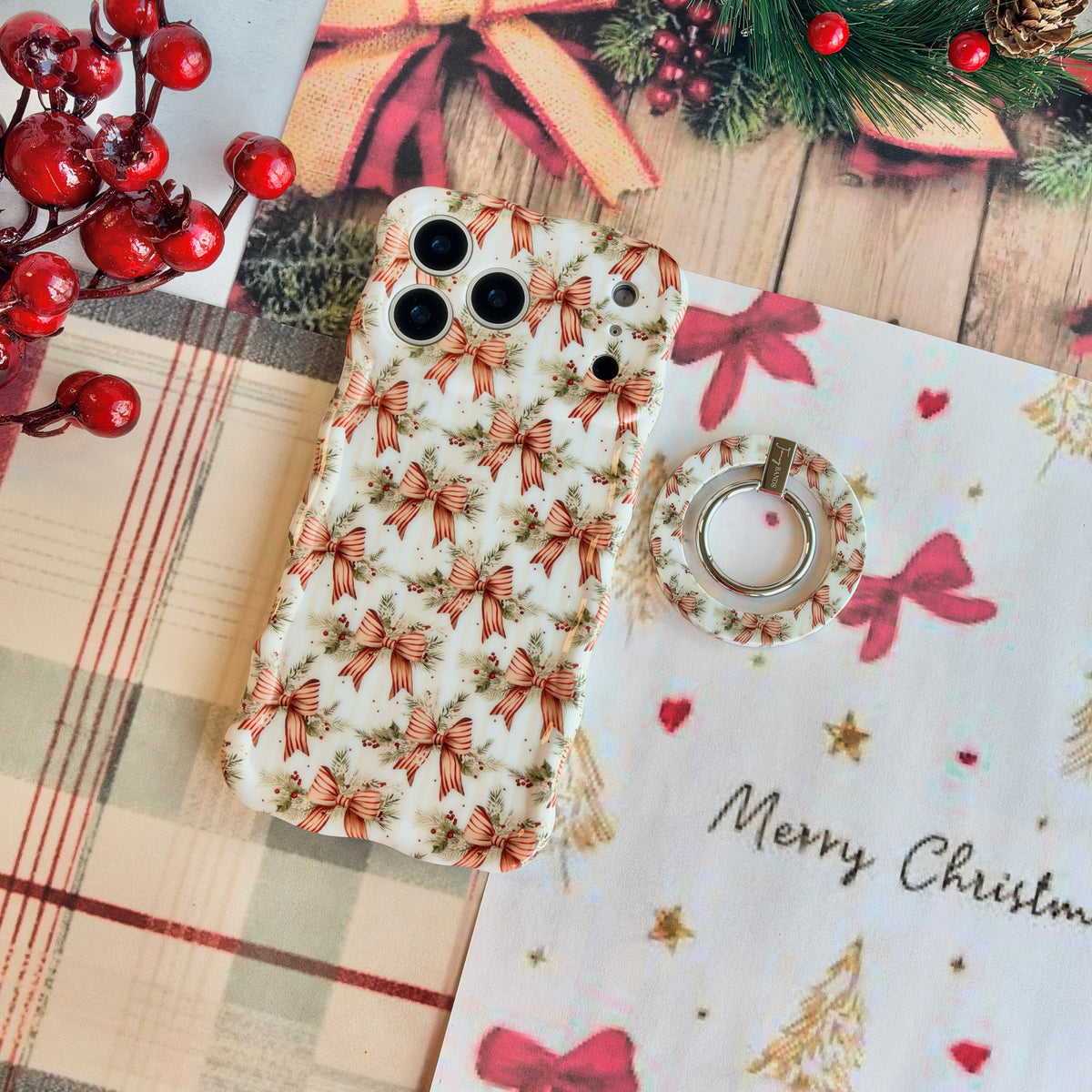 Deck Yourself in Bows & Holly MagSafe grip ring lying beside the matching iPhone case on cream plaid with red berries and Merry Christmas paper