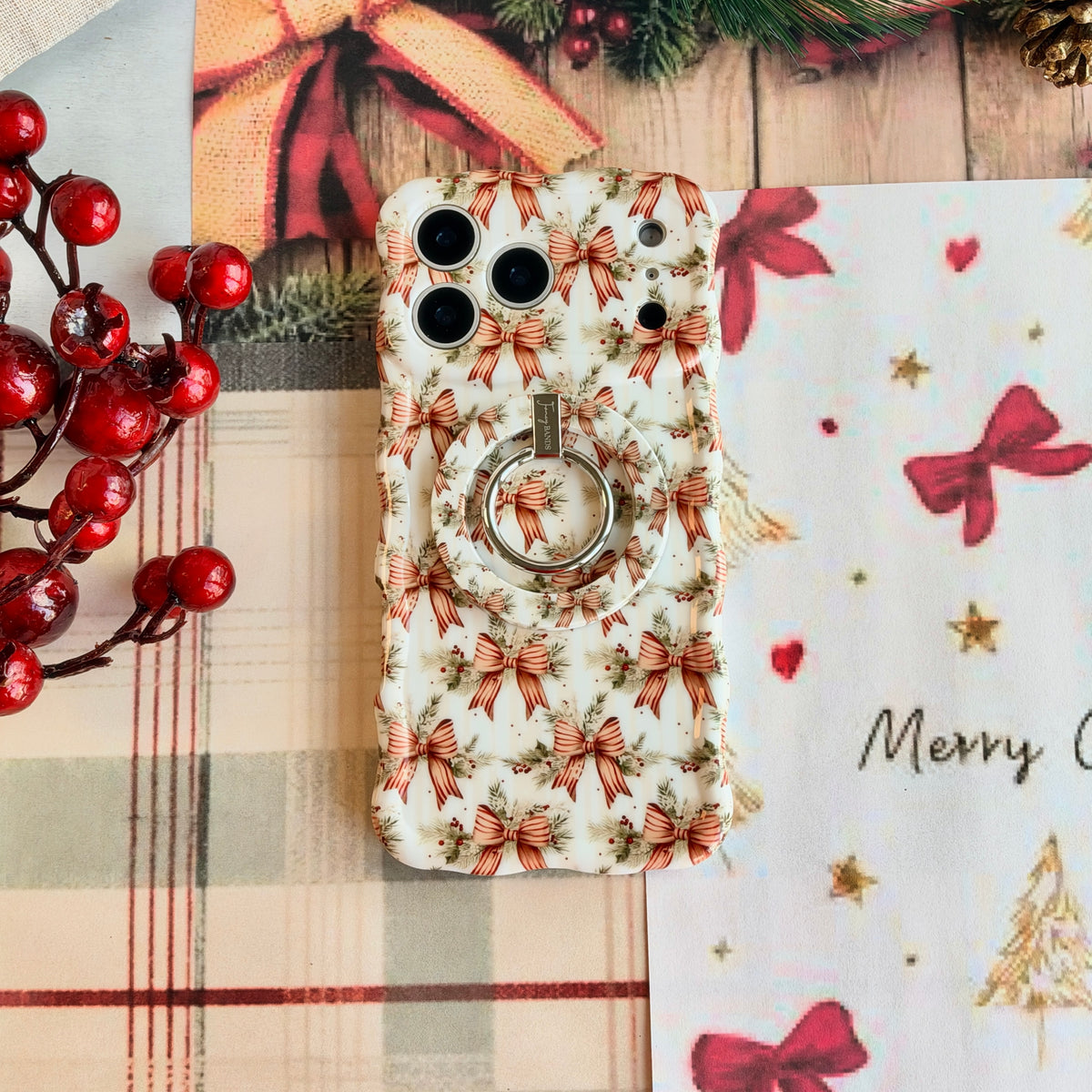 Close-up of the Deck Yourself in Bows & Holly MagSafe grip ring on the matching bow-print iPhone case, on cream plaid with red berries
