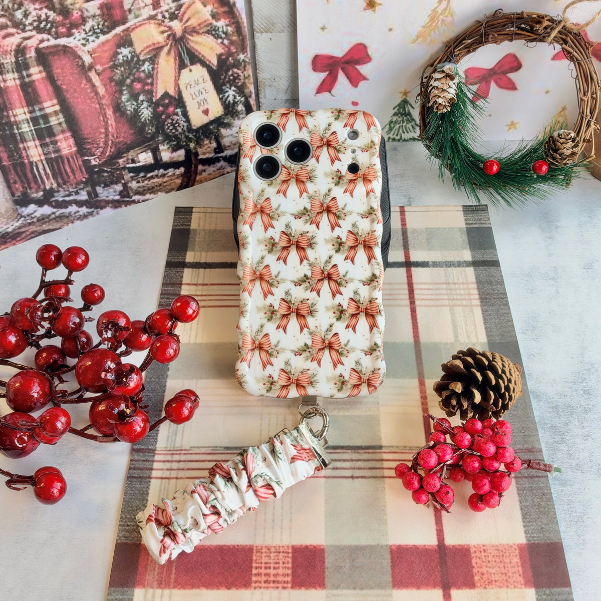 Flat lay of the Deck Yourself in Bows & Holly iPhone case with its bow-print scrunchie wrist strap, a pine wreath, pinecone and red berries