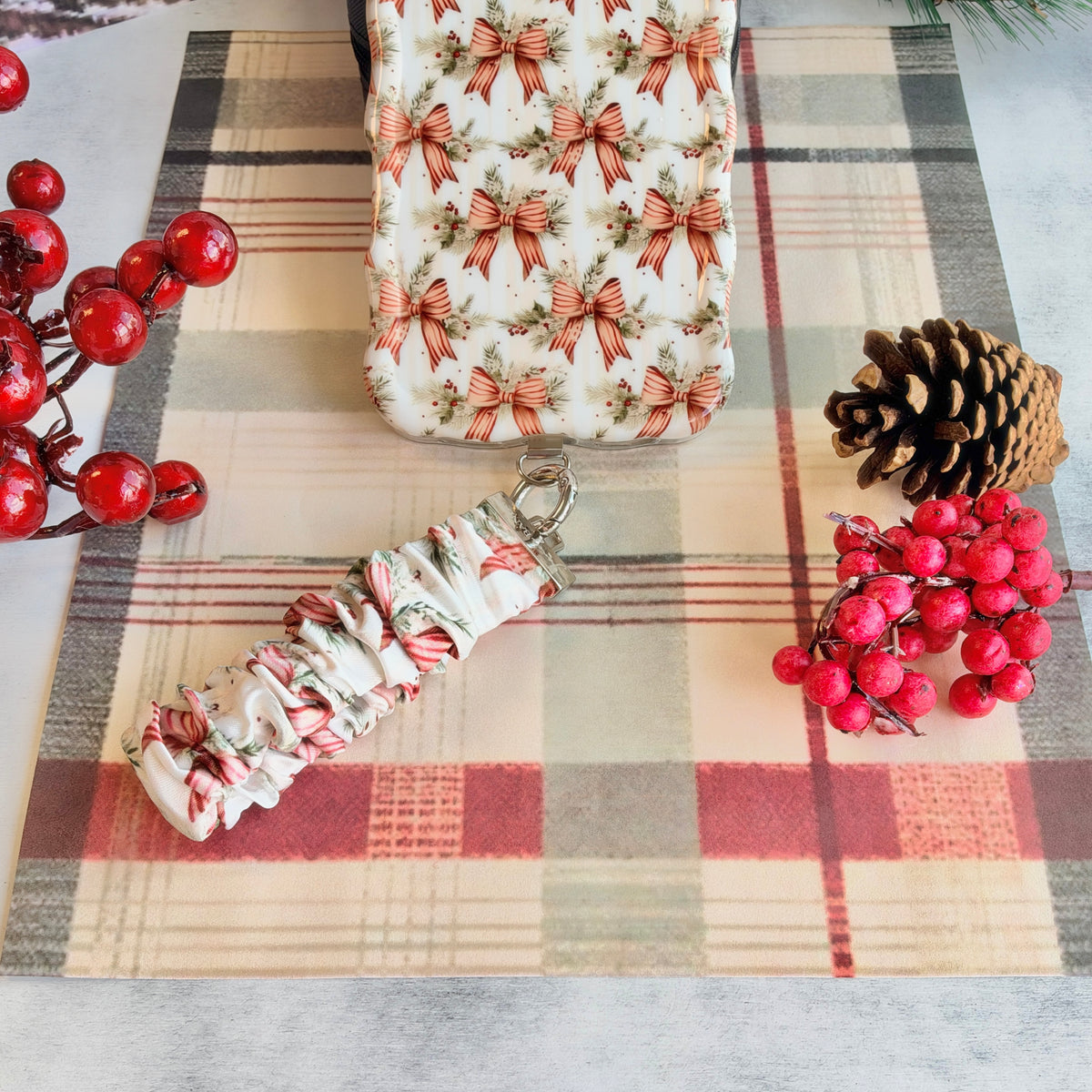 Deck Yourself in Bows & Holly scrunchie phone wrist strap clipped below the matching iPhone case on cream plaid, with a pinecone and red berries