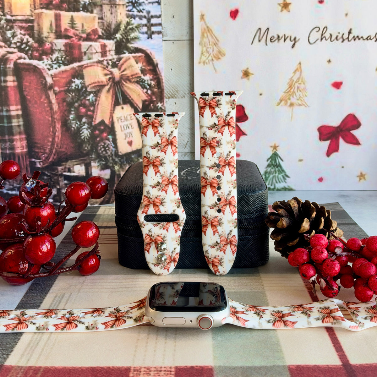 Deck Yourself in Bows & Holly Apple Watch band shown on an Apple Watch and as two straps on a black jewelry box, red ribbon bows and holly on ivory