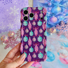 Hand holding the Just One More Ornament Wavy Luxe MagSafe iPhone case, pink and aqua watercolor Christmas ornaments on plum, in front of lavender snowflakes and an aqua tinsel tree