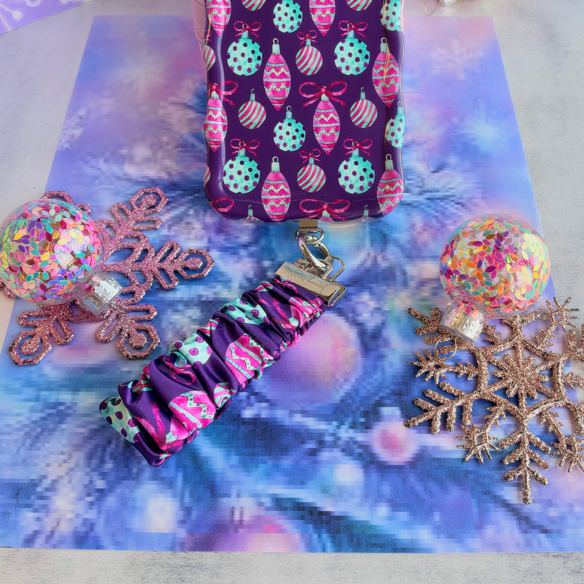 Just One More Ornament scrunchie phone wrist strap clipped below the matching iPhone case on aqua tinsel, with pink glitter snowflakes and confetti ornaments