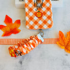 An orange gingham Happy Fall Y'all scrunchie phone wrist strap with a jack-o'-lantern and 'happy fall y'all' print, clipped to the matching Happy Fall Y'all Wavy Luxe iPhone case, styled with autumn maple leaves.