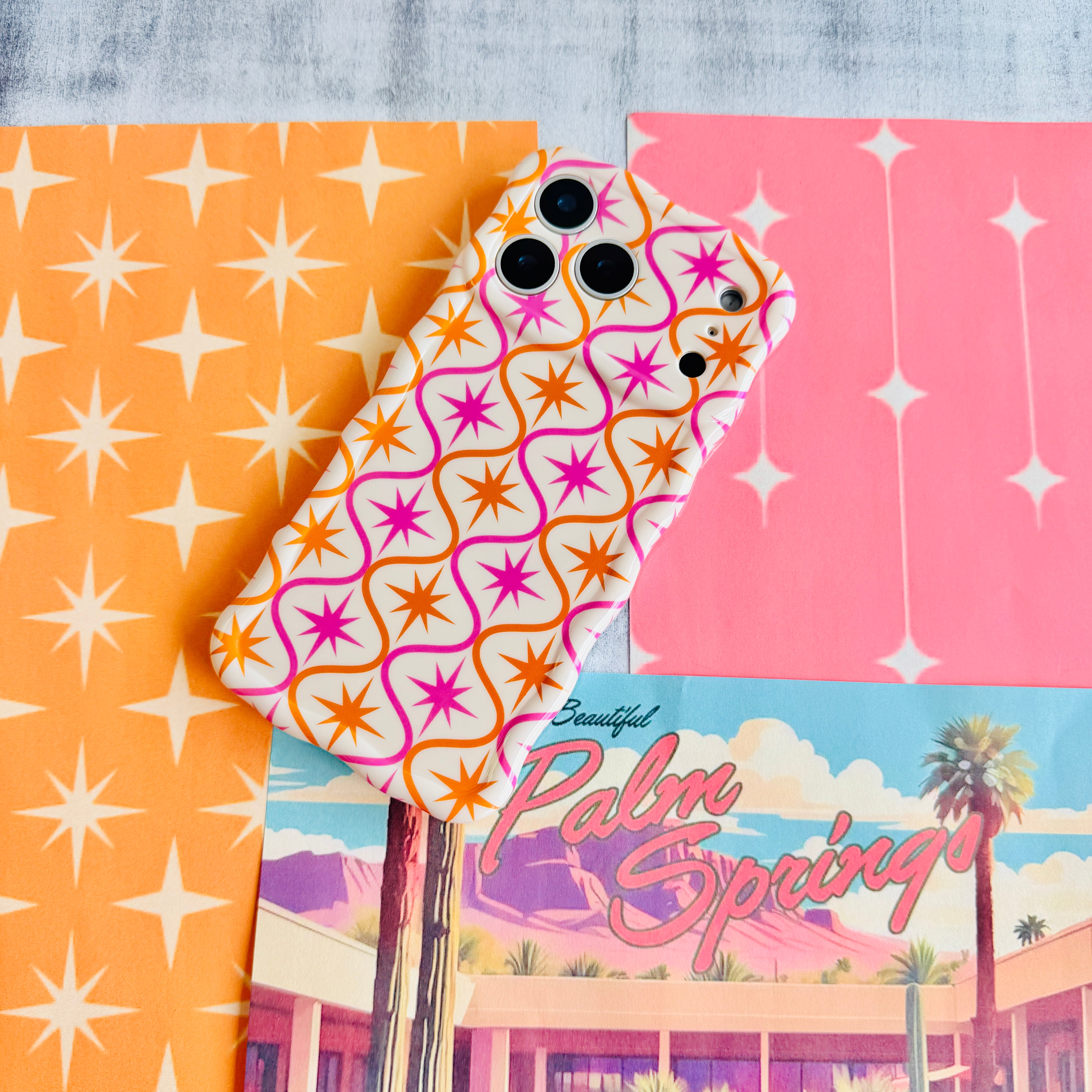Trendy wavy iPhone case featuring a pink and orange starburst geometric pattern on a cream background with a high-gloss finish.