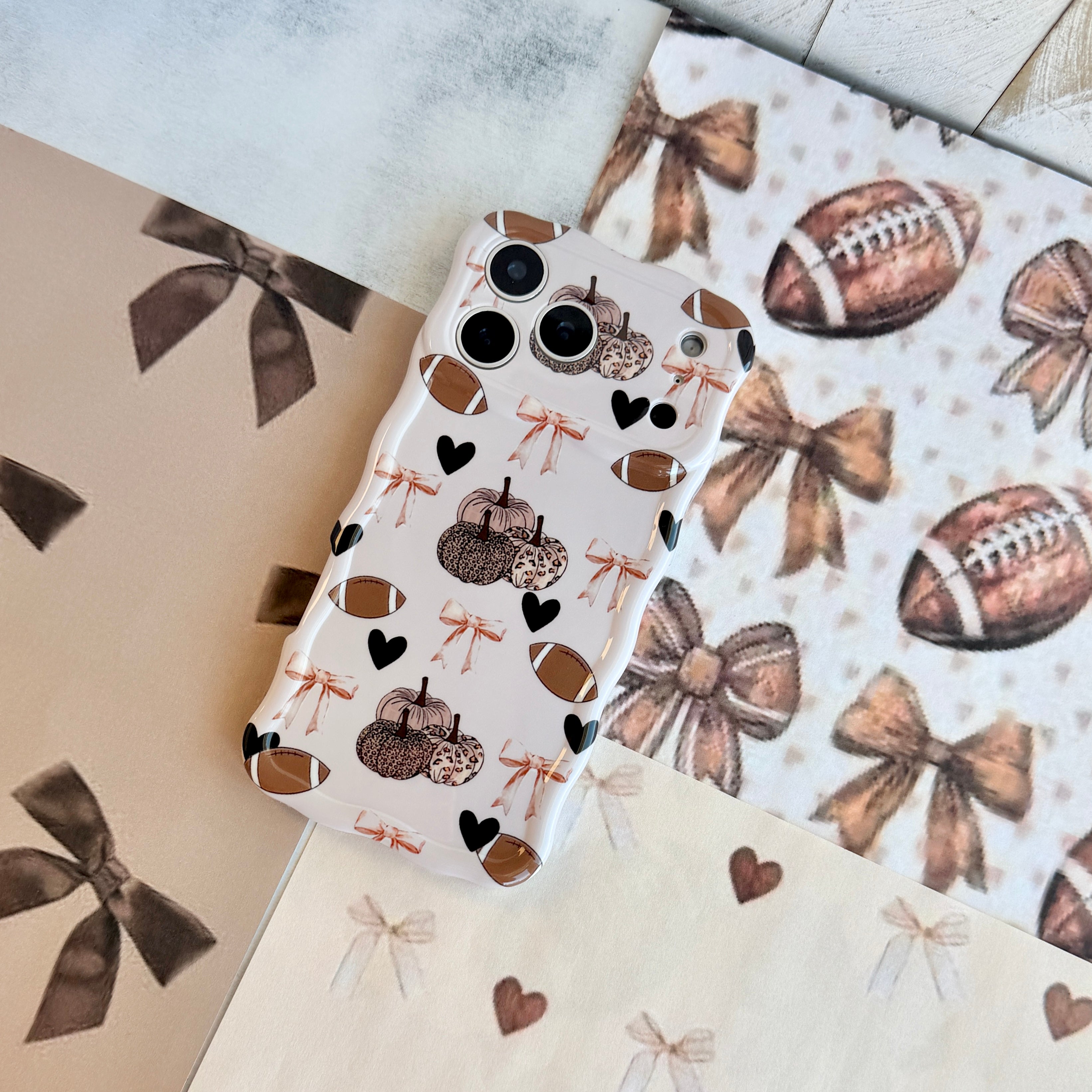 Game Day Darling Wavy Luxe MagSafe iPhone case with a cream coquette print of footballs, leopard pumpkins, bows and hearts from Fancy Bands