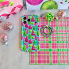 Merry & Bright, Totally! MagSafe grip ring lying beside the matching iPhone case on pink and green plaid with a lime gift bow