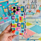 Hand holding the Mod Mood Wavy Luxe MagSafe iPhone case with teal, lime, pink and navy rounded squares on a retro atomic starburst backdrop