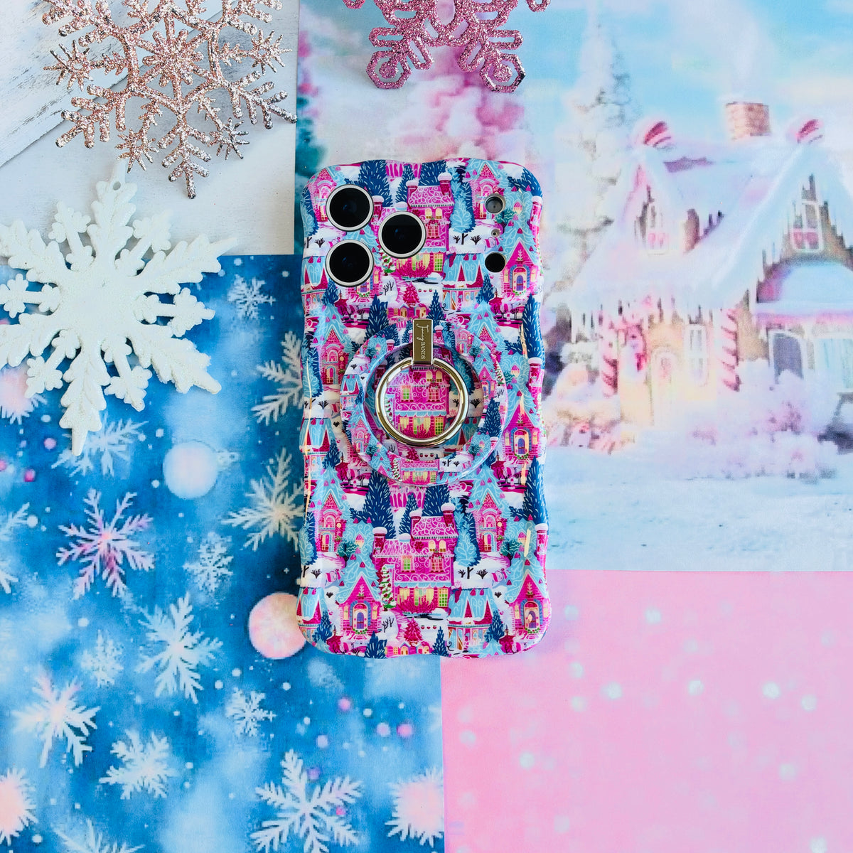Sugarplum Square MagSafe grip ring snapped onto the matching Christmas village iPhone case, on blue snowflake paper with glitter snowflakes