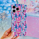 Hand holding the Sugarplum Square Wavy Luxe MagSafe iPhone case, pink gingerbread houses and blue pines in the snow, in front of a snowy pastel village backdrop