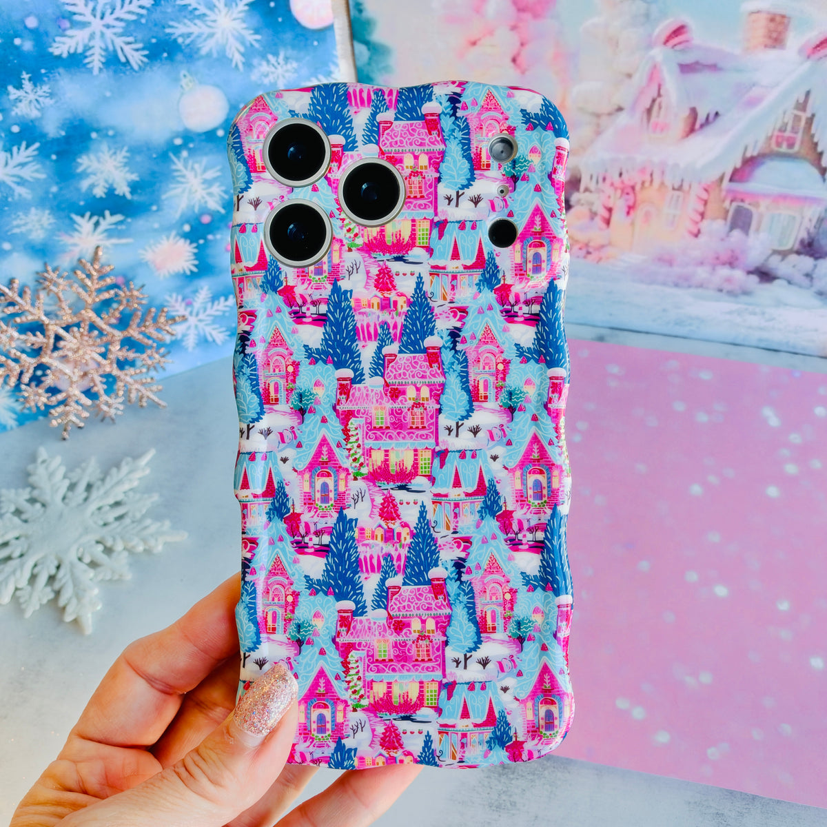Hand holding the Sugarplum Square Wavy Luxe MagSafe iPhone case, pink gingerbread houses and blue pines in the snow, in front of a snowy pastel village backdrop