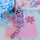 Sugarplum Square iPhone case with its Christmas village scrunchie wrist strap on pink and blue snowflake paper, beside a pink bottle-brush tree