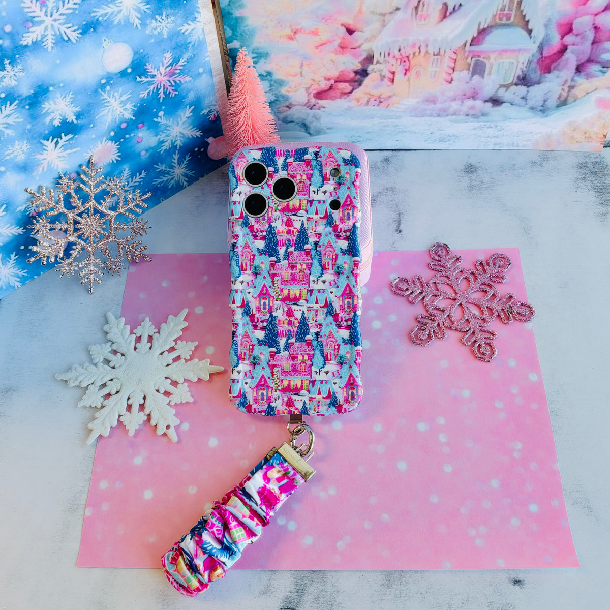 Sugarplum Square iPhone case with its Christmas village scrunchie wrist strap on pink and blue snowflake paper, beside a pink bottle-brush tree