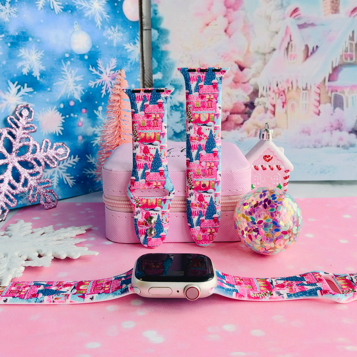 Sugarplum Square Apple Watch band shown on an Apple Watch and as two straps on a pink jewelry box, pink gingerbread houses and blue pines