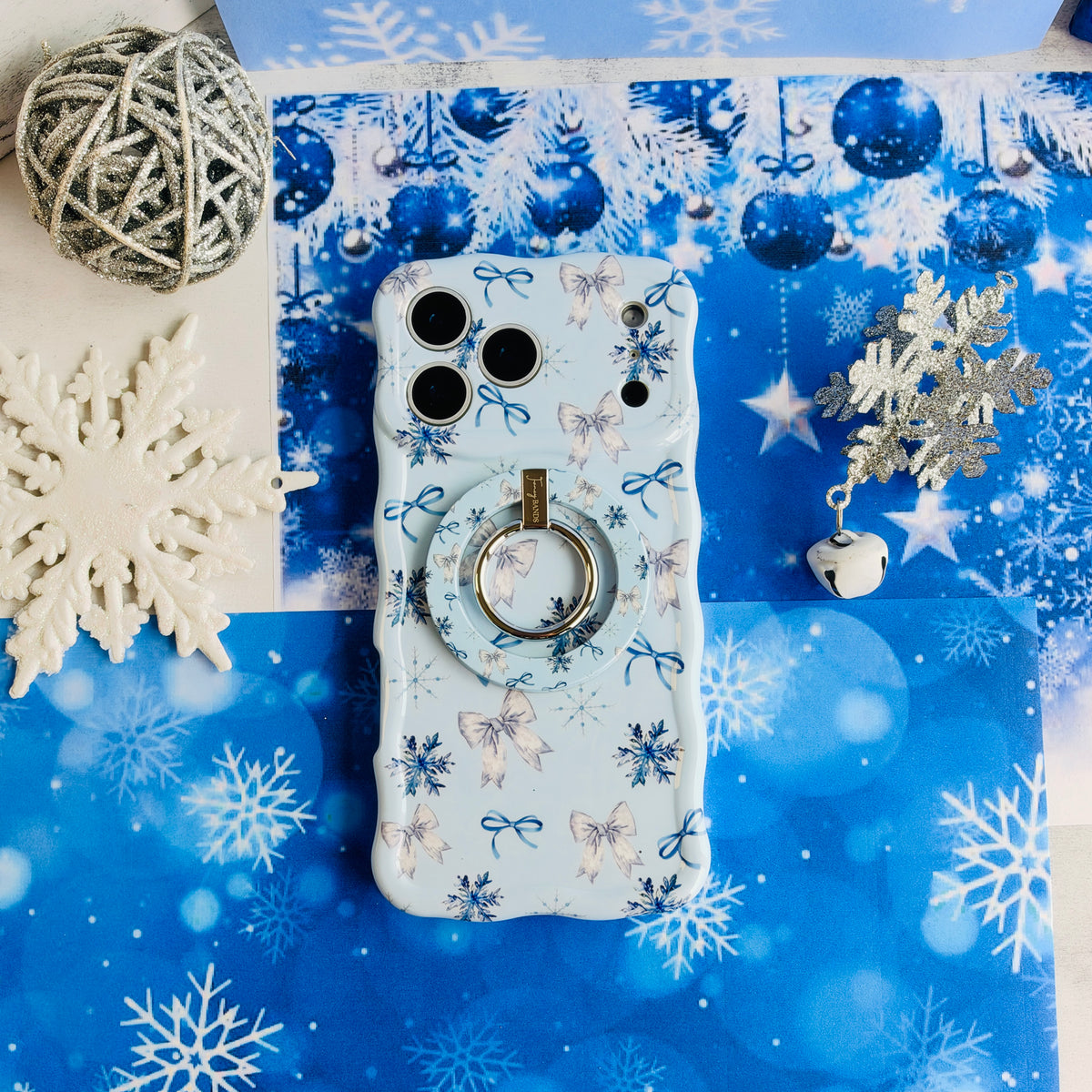 Snow Pretty In Blue MagSafe grip ring snapped onto the matching snowflake bow iPhone case, with a jingle bell and silver snowflakes on blue paper