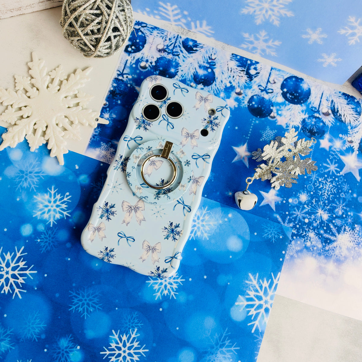 Snow Pretty In Blue iPhone case with the matching grip ring snapped on, beside a white snowflake and a silver tinsel ball on blue paper