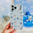 Hand holding the Snow Pretty In Blue Wavy Luxe MagSafe iPhone case, silver bows and navy snowflakes on powder blue, over blue snowflake paper