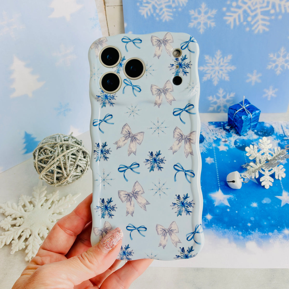 Hand holding the Snow Pretty In Blue Wavy Luxe MagSafe iPhone case, silver bows and navy snowflakes on powder blue, over blue snowflake paper