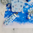Snow Pretty In Blue scrunchie phone wrist strap clipped below the matching iPhone case on blue snowflake paper with glitter snowflakes