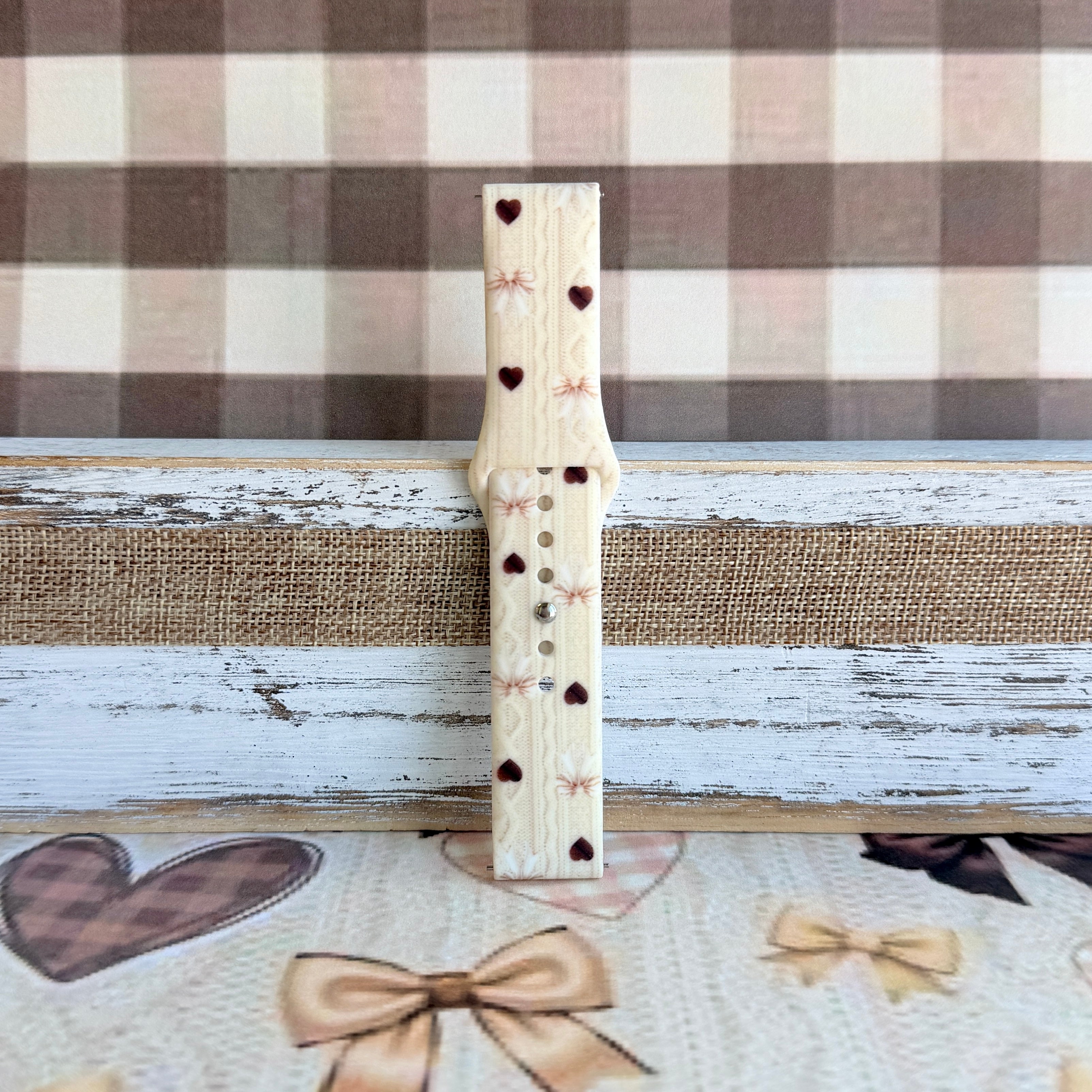 Close-up detail of Sweater Weather Sweetheart Samsung Galaxy Watch band showing the cream cable-knit texture, taupe bow print, and brown heart accents.