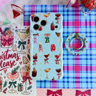 Holiday Spirits, Please? MagSafe grip ring lying beside the matching iPhone case on pink and blue plaid with a Christmas, please print