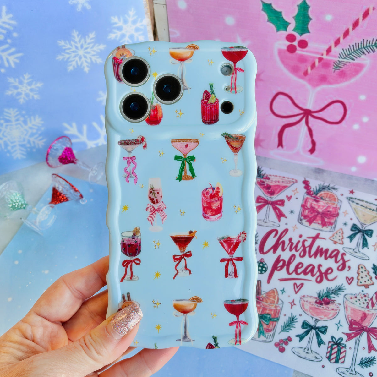 Hand holding the Holiday Spirits, Please? Wavy Luxe MagSafe iPhone case, holiday cocktails and bows on powder blue, over pink cocktail and bow paper